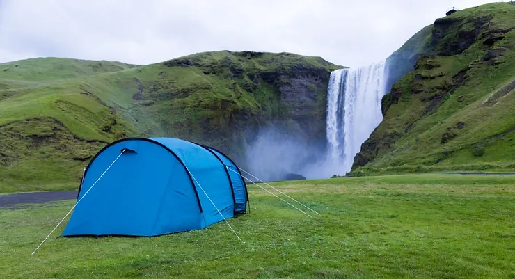 A blue tent set up on a grassy area near a powerful waterfall in Iceland, with lush green hills surrounding the scene.