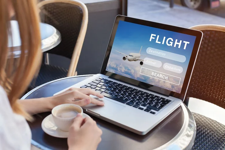 A person sitting at a cafe table with a laptop open to a flight booking website, displaying options to search for destination, arrival, and departure. The person holds a cup of coffee in one hand while using the laptop with the other hand.