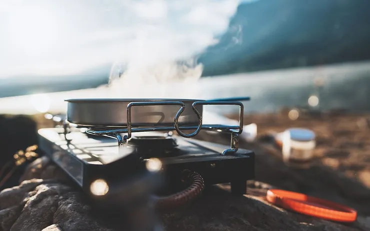 A close-up of a portable camping stove with a pan, set up on a rocky surface with steam rising from it, against a scenic background of a lake and mountains in Iceland.