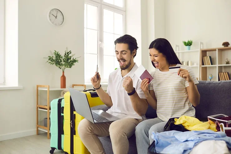 A cheerful couple sitting on a couch, excitedly using a laptop to book travel deals. The man holds a credit card, and the woman holds a passport and another card. Luggage and packed clothes are visible in the background, indicating preparation for travel.