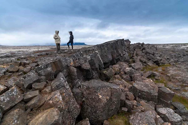 Two people stand on a rocky landscape near Surtshellir Cave in Iceland, surrounded by rugged terrain and a cloudy sky.