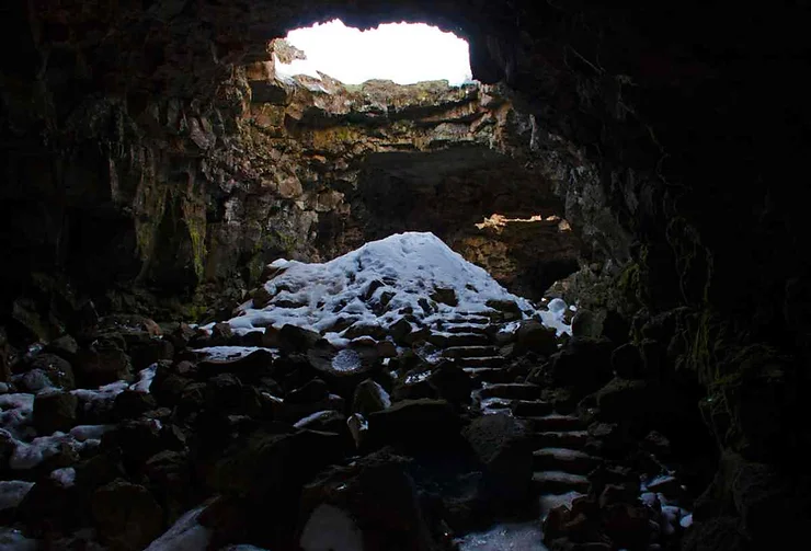 Interior view of Surtshellir Lava Cave in Iceland, showing a rocky floor with patches of snow and a rugged cave opening leading to daylight.