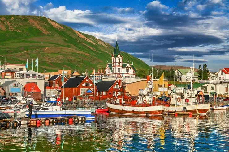 A vibrant view of Húsavík, Iceland, with its colorful buildings, traditional fishing boats, and the picturesque wooden church, all set against a backdrop of lush green hills and a bright blue sky.