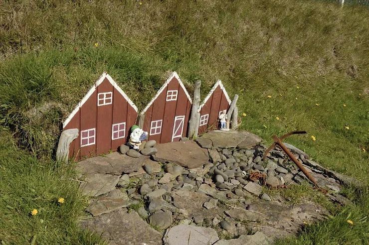 A set of small red elf houses with white trim nestled in the grass, with small elf figurines placed outside, on a sunny day in Iceland.