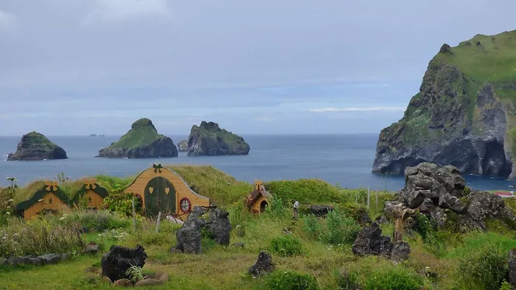 A scenic view of a grassy area with small, charming elf houses in Iceland, set against the backdrop of the ocean and rocky islands.