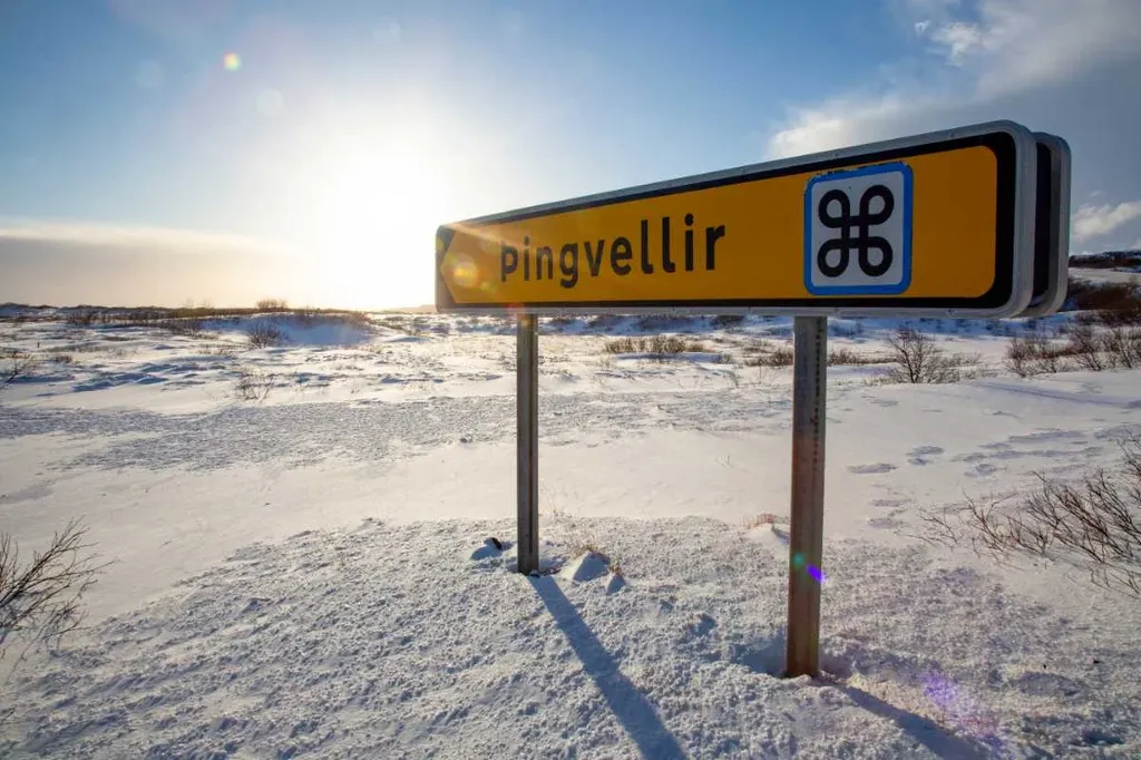 Signpost pointing to Thingvellir in Iceland, standing in a snow-covered landscape with the sun rising or setting in the background.