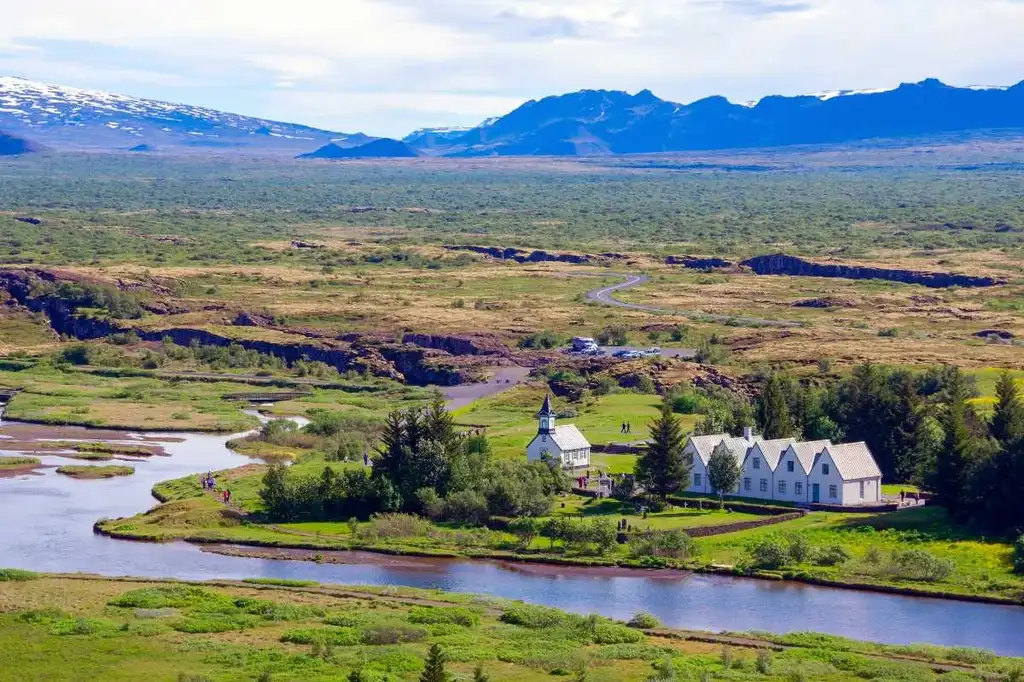 Aerial view of Thingvellir National Park in Iceland, featuring a cluster of white buildings including Thingvellir Church, surrounded by lush greenery, a winding river, and distant mountains.