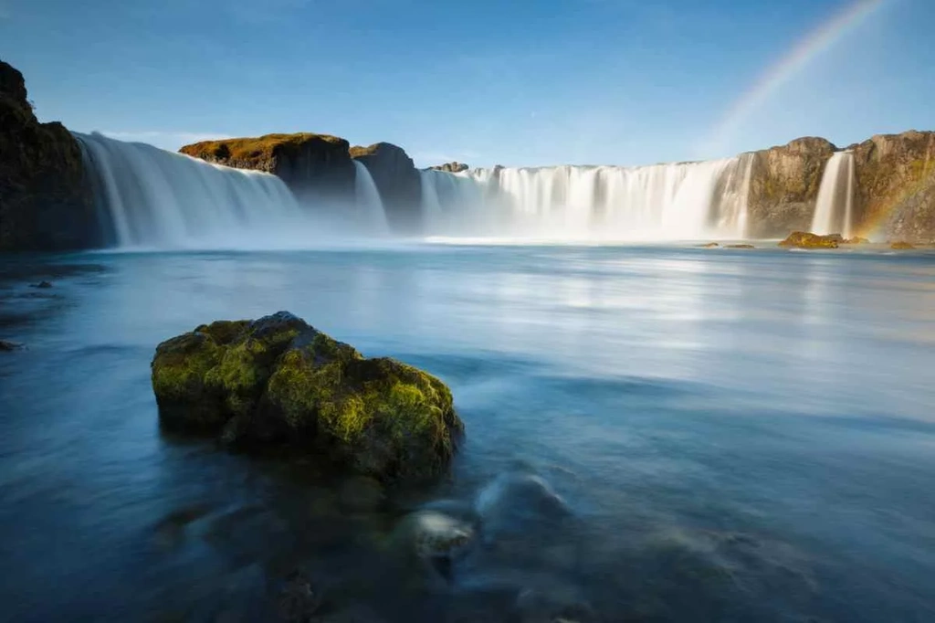 Long-exposure shot of Goðafoss waterfall in Iceland, with smooth flowing water, a moss-covered rock in the foreground, and a faint rainbow in the sky