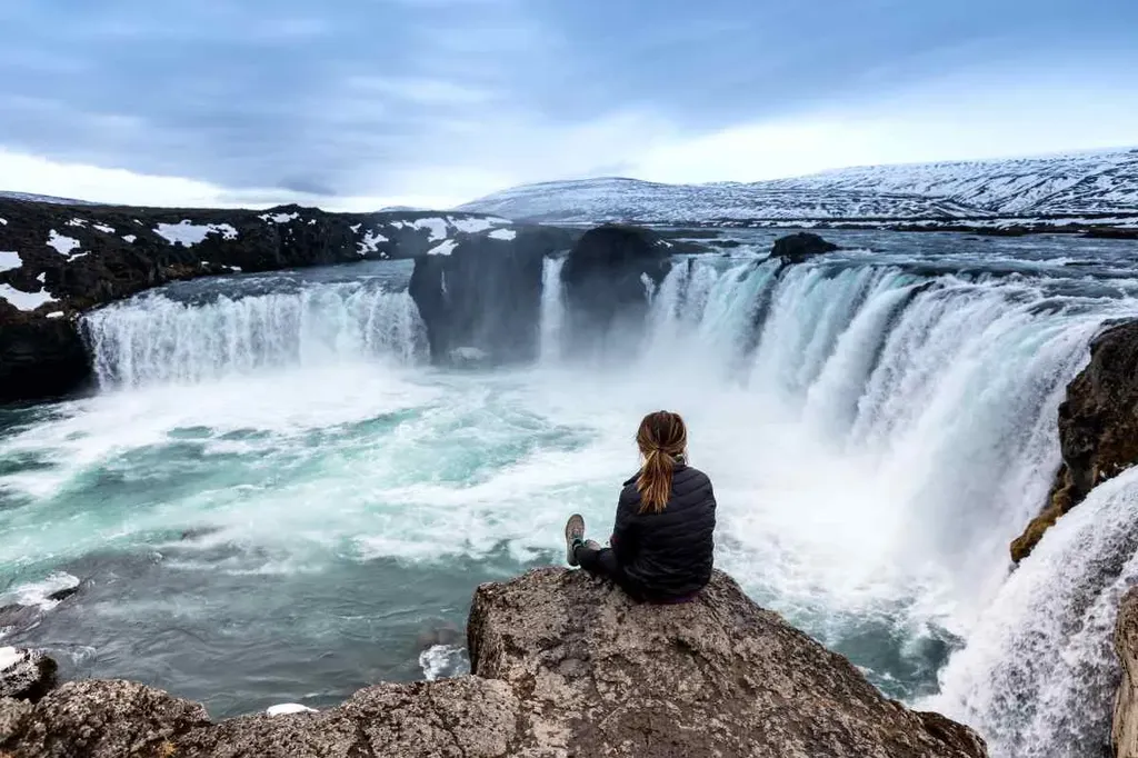 Woman sitting on a cliff edge overlooking the powerful Goðafoss waterfall in Iceland, surrounded by snow-covered mountains and mist from the falls