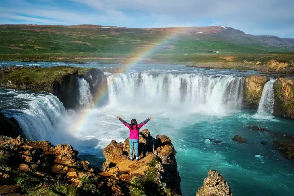 Woman standing on a cliff with arms raised, overlooking Goðafoss waterfall in Iceland, with a vibrant rainbow arching across the falls and lush green landscape