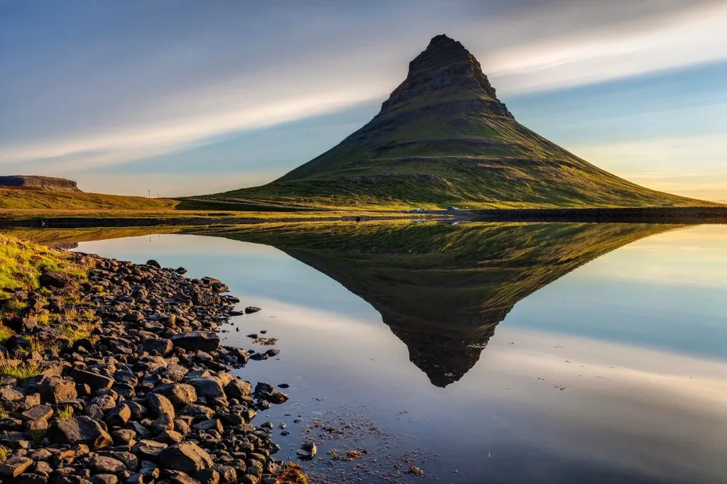 Kirkjufell mountain in Iceland perfectly reflected in the calm waters of a nearby lake, with rocky shoreline and soft sunlight illuminating the scene