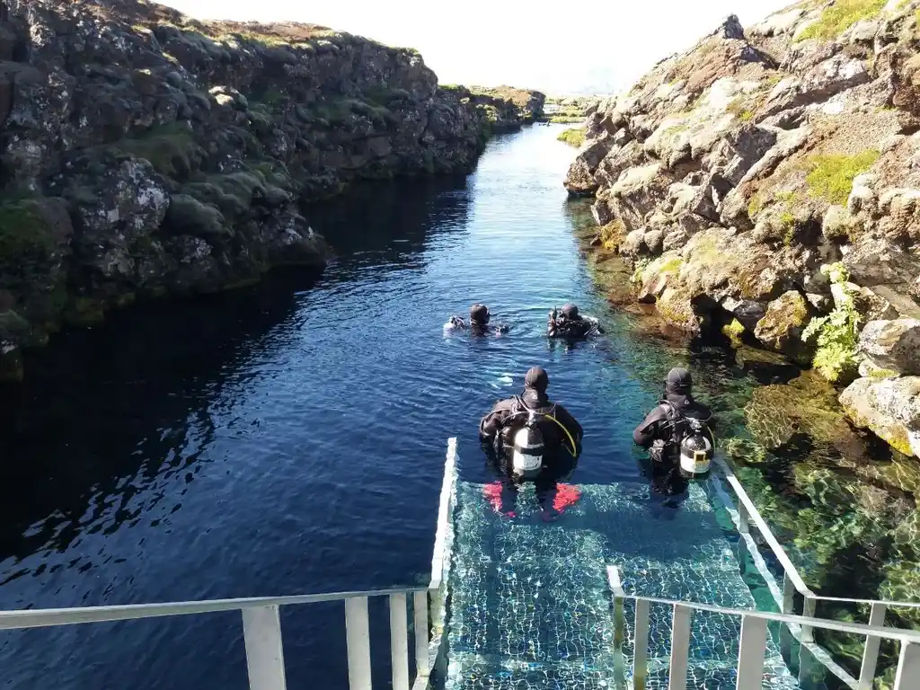 Group of divers preparing to enter the crystal-clear waters of the Silfra Fissure in Thingvellir National Park, Iceland, surrounded by rocky cliffs