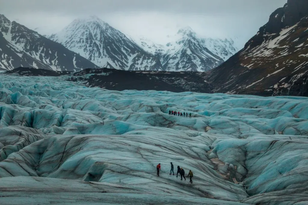 Group of hikers trekking across the blue ice of a glacier in Vatnajökull National Park, Iceland, with snow-capped mountains looming in the background