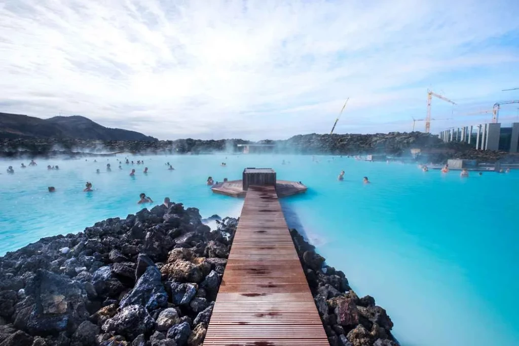 A view of the Blue Lagoon in Iceland, featuring a wooden boardwalk leading into the steaming, milky-blue geothermal waters with people enjoying a soak amidst the surrounding volcanic rocks.