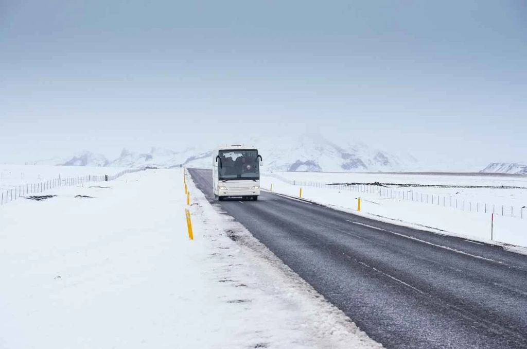 A white bus driving on a snow-covered road in Iceland, surrounded by vast snowy landscapes and distant mountains