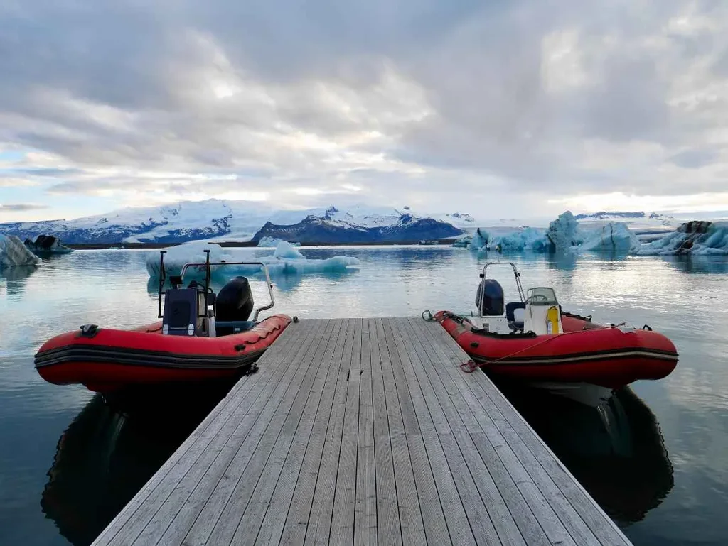 Two red inflatable boats docked on a wooden pier at Jökulsárlón Glacier Lagoon in Iceland, with floating icebergs and snow-capped mountains in the background under a cloudy sky.