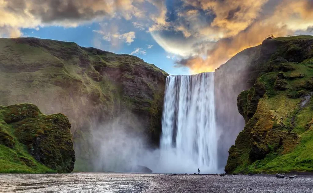 Skogafoss Waterfall in Iceland, with a dramatic cascade of water plunging down a steep cliff surrounded by lush green hills under a vibrant sky filled with orange and blue hues. A small figure stands near the base of the waterfall, highlighting the immense scale of the landscape.