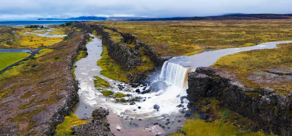 Aerial view of Thingvellir National Park in Iceland, showing a waterfall cascading over a cliff into a river surrounded by rugged terrain with scattered patches of greenery under a cloudy sky.