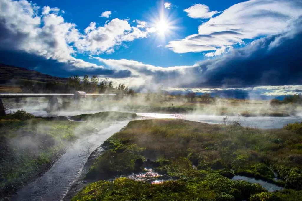 Scenic view of Deildartunguhver hot springs in Iceland with steam rising from the water under a bright sun and dramatic clouds.