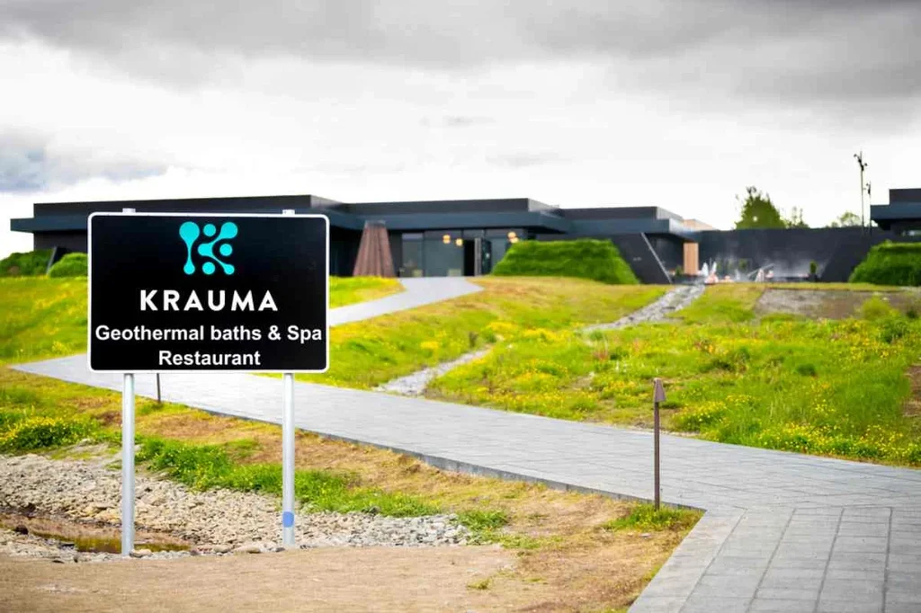 Krauma Geothermal Baths & Spa entrance with a sign highlighting the restaurant, set against the modern building and lush greenery in Iceland.