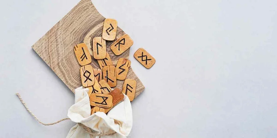 Set of wooden rune tiles spilling out from a cloth pouch onto a wooden board, used for Norse divination and spiritual guidance.