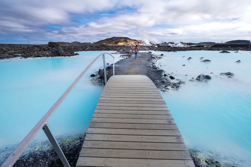 Scenic view of the Blue Lagoon in Iceland with a wooden bridge crossing the vibrant blue geothermal waters, surrounded by rugged volcanic landscape.