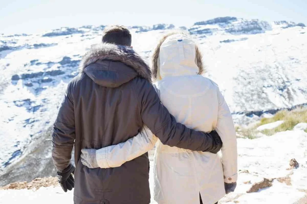 Couple in winter coats embracing while standing on a snowy mountainside in Iceland, enjoying a scenic winter landscape.