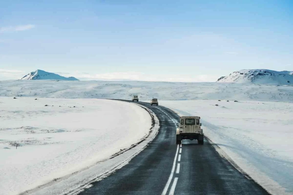 Vehicles driving on a snowy road in Iceland, surrounded by a vast, snow-covered landscape with distant mountains under a clear blue sky.