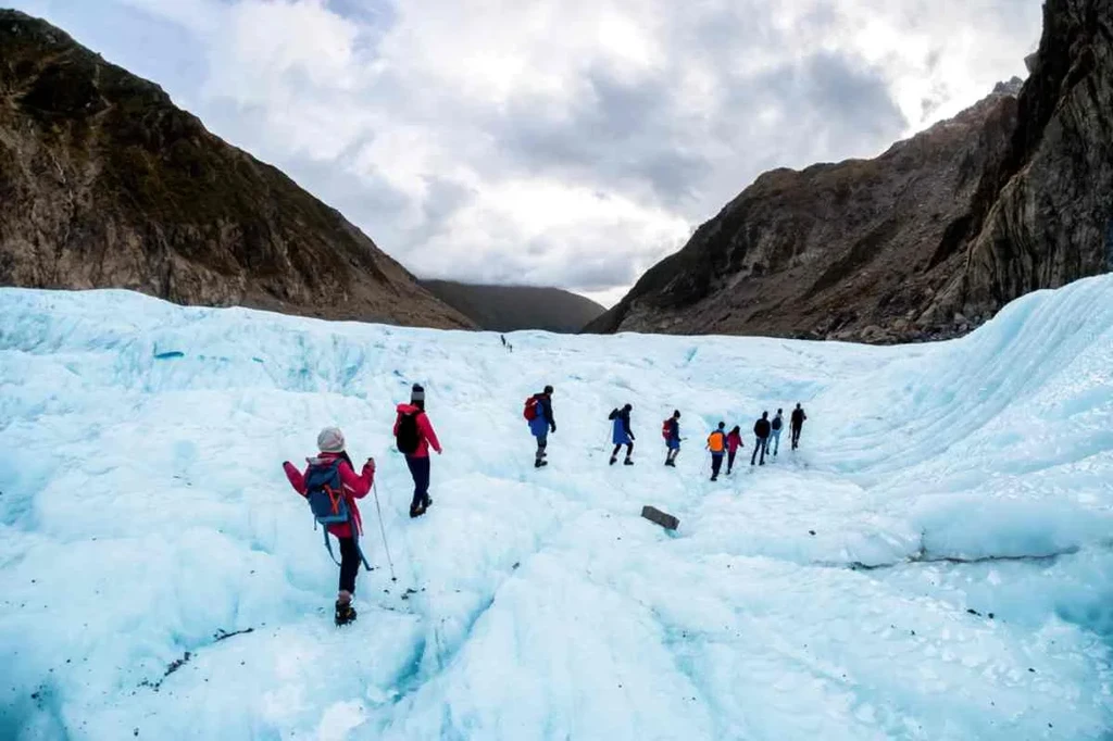 Group of hikers equipped with winter gear trekking across a blue glacier in Iceland, surrounded by rugged mountains under a cloudy sky.