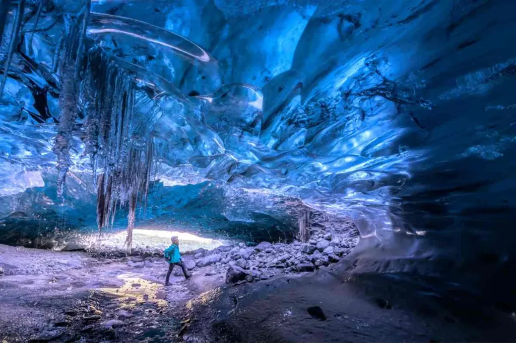 Person exploring a mesmerizing blue ice cave in Iceland, with intricate ice formations and icicles hanging from the ceiling, illuminated by natural light.