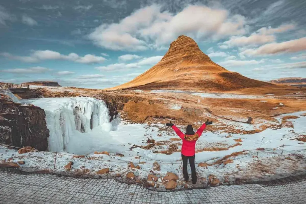 Traveler in a red jacket standing in front of the iconic Kirkjufell mountain and frozen waterfall in Iceland during winter, arms raised in awe.