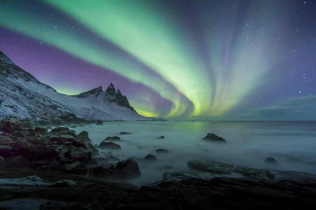 Stunning Northern Lights illuminating the night sky over a rugged coastline in Iceland, with snow-covered peaks and rocky shores.