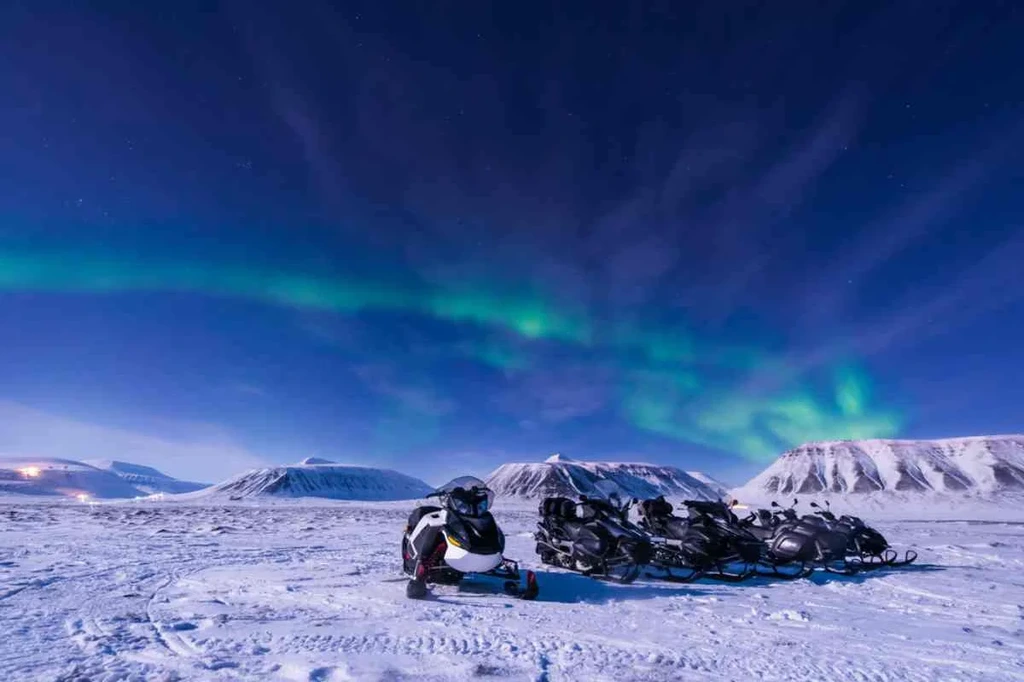 Snowmobiles parked on a snowy landscape in Iceland, under a vibrant display of the Northern Lights, with snow-covered mountains in the background.