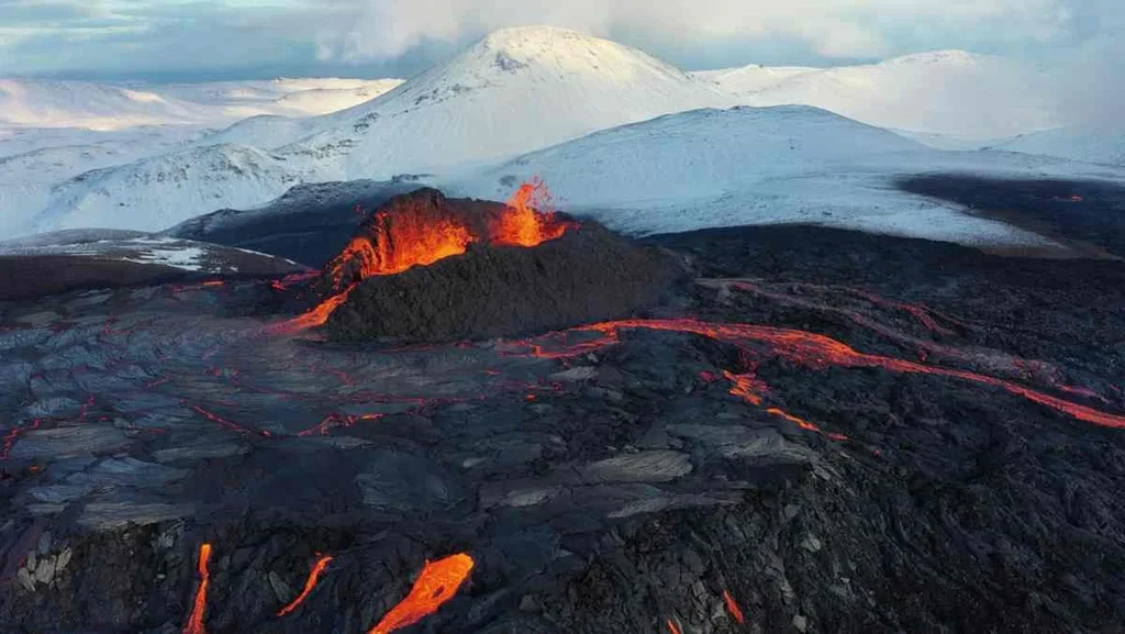 Active volcano in Iceland with bright red lava flowing over dark volcanic rock, set against a backdrop of snow-covered mountains.