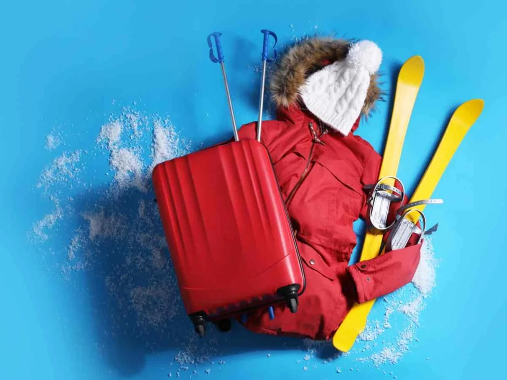 Winter travel essentials laid out on a blue background, including a red suitcase, ski gear, a red parka, and a white knit beanie with snow scattered around.