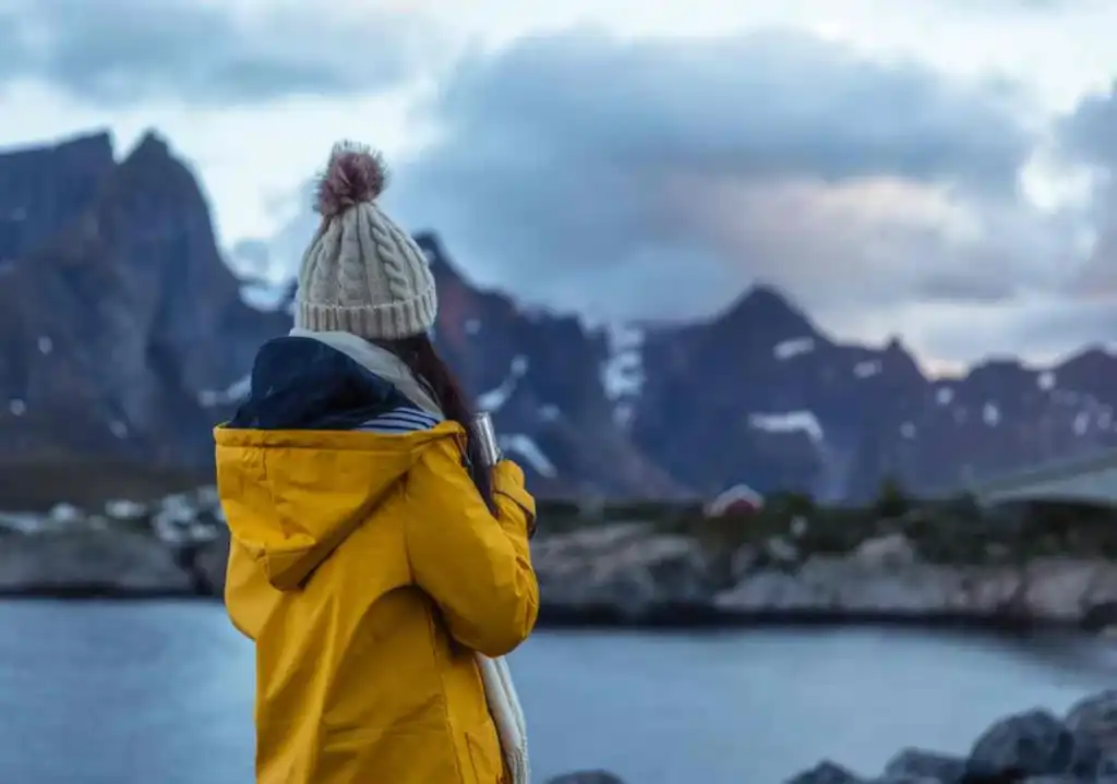 Person in a yellow jacket and knit beanie standing by a fjord in Iceland, gazing at the snow-capped mountains under a cloudy sky.