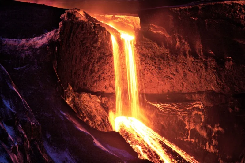 Molten lava cascading down a rocky cliff during a volcanic eruption, creating a glowing waterfall of fire against the dark landscape.