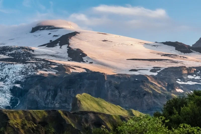 Snow-covered mountain peak illuminated by the sunlight, with lush green vegetation in the foreground under a partly cloudy sky.