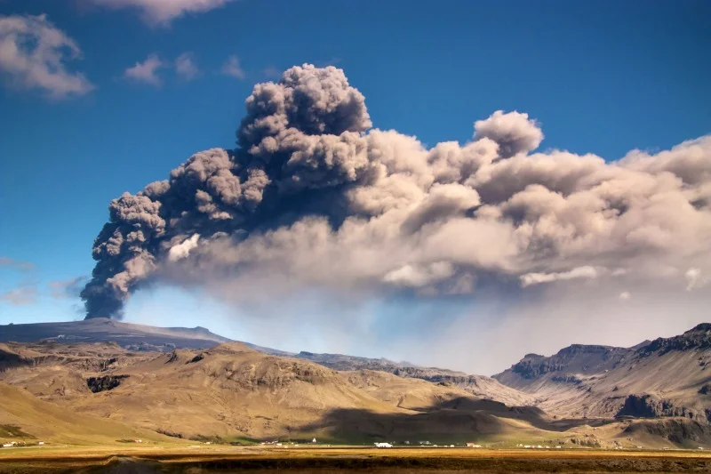 Massive plume of ash and smoke rising from a volcanic eruption, contrasting against a clear blue sky with rugged mountains in the background.