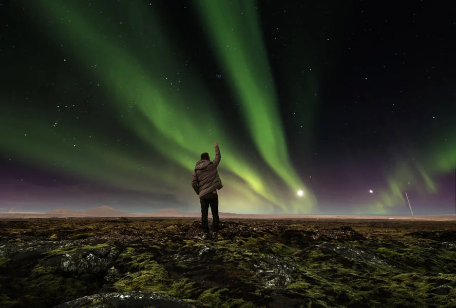 Person standing on a rocky terrain under a vibrant display of the Northern Lights, reaching towards the illuminated night sky.