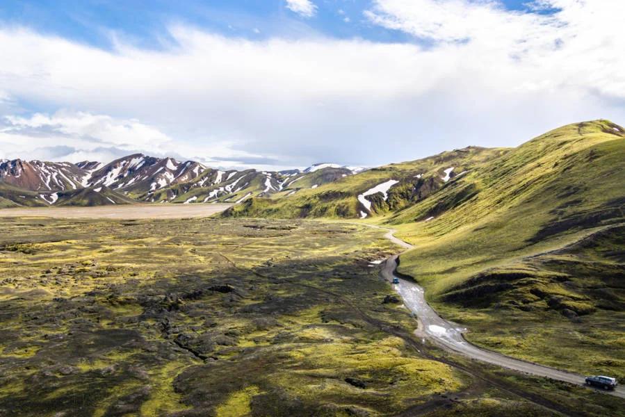 Expansive view of a rugged landscape with rolling green hills and snow-capped mountains under a partly cloudy sky, featuring a winding dirt road.