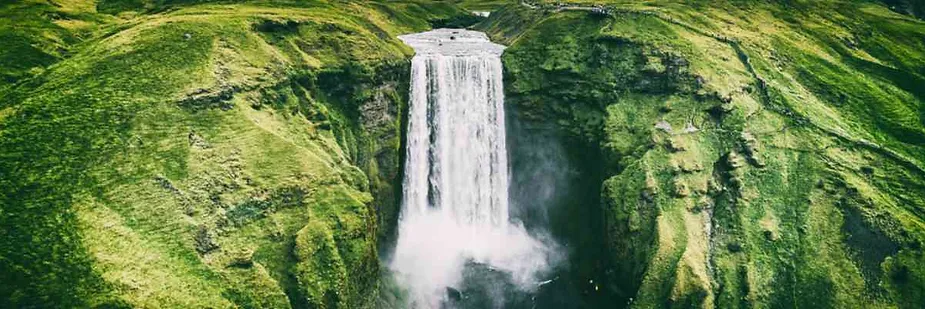 Aerial view of Skógafoss waterfall in Iceland cascading down a lush green cliff, surrounded by vibrant moss-covered landscapes.