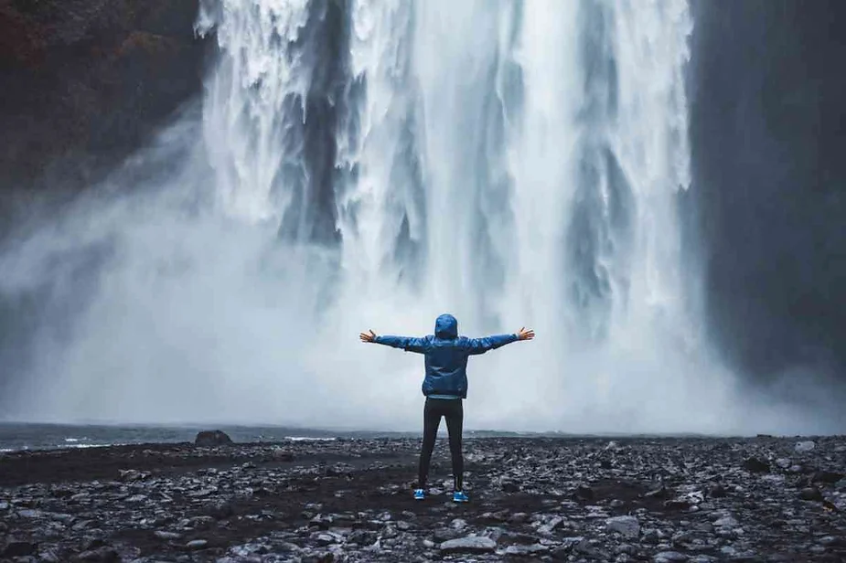 Traveler in a blue jacket standing with arms wide open in front of the powerful Skógafoss waterfall in Iceland, embracing the mist and raw energy of nature.