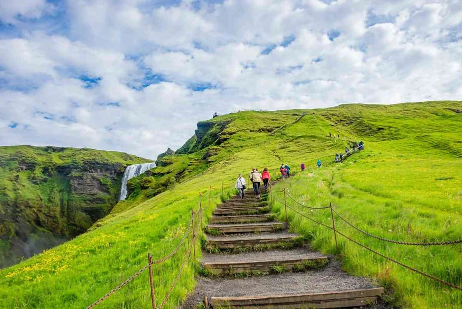 Hikers climbing wooden stairs surrounded by lush green hills, approaching the top of Skógafoss waterfall in Iceland under a cloudy sky.