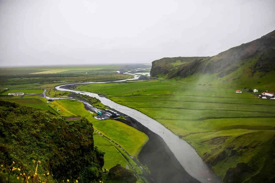 Panoramic view of the winding river surrounded by lush green fields and hills from the top of Skógafoss waterfall in Iceland on a foggy day.