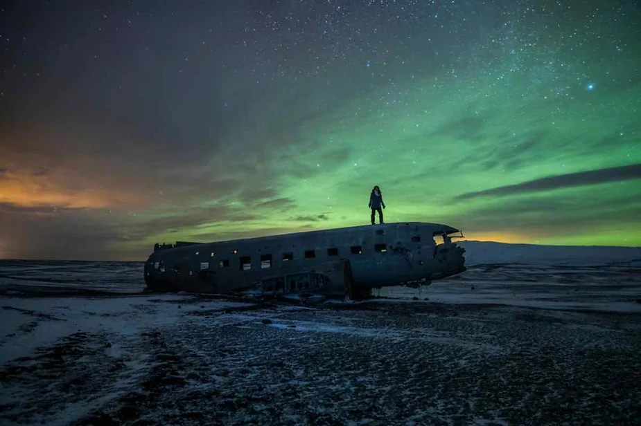 Person standing on the wreckage of a plane under the stunning display of the Northern Lights in Iceland, with a starry sky and snowy landscape.