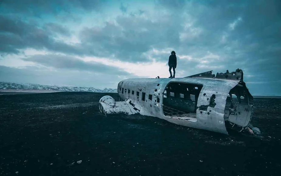 Person standing on the wreckage of an abandoned plane on a desolate black sand beach in Iceland, under a cloudy sky.