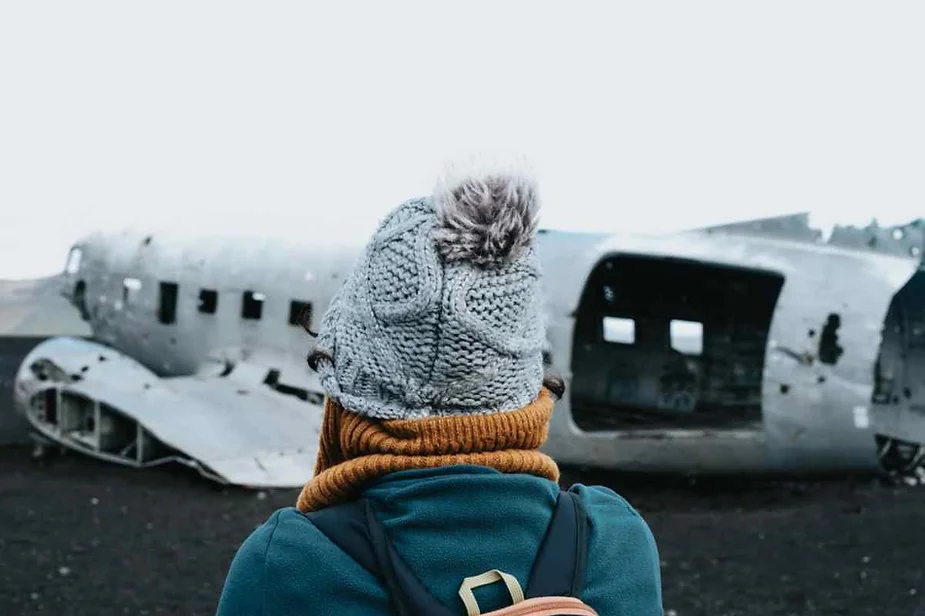 Traveler wearing a winter hat and scarf standing in front of the iconic plane wreck on Iceland's black sand beach, gazing at the ruins.