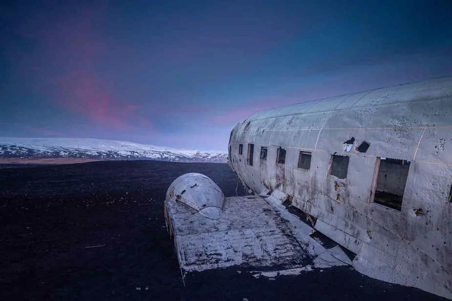 Abandoned DC-3 plane wreck on Iceland's black sand beach under a dramatic twilight sky, with distant snow-capped mountains in the background.