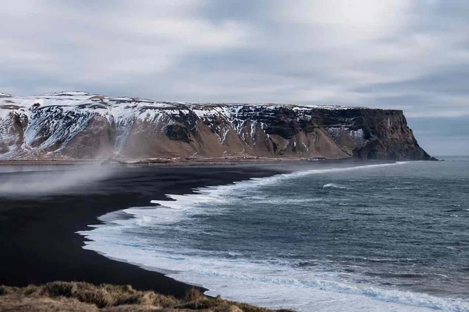 Scenic view of Iceland's black sand beach with waves crashing against the shore, surrounded by snow-covered cliffs under a cloudy sky.